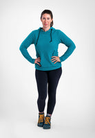 A woman modeling a teal Highlander hoodie with black drawstrings, paired with black leggings and hiking boots, standing against a light gray studio background.