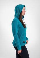 A woman in profile wearing a teal Highlander Hoodie and black leggings, standing against a light gray studio background.