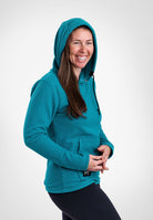 A woman wearing a teal Highlander Hoodie and black leggings, smiling and posing against a light gray studio background.