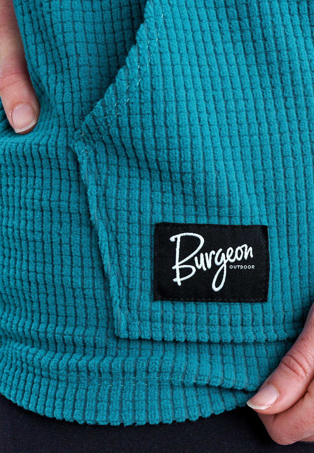Close-up of a teal Highlander Hoodie with a black sewn-on brand patch reading 'Burgeon OUTDOOR' held by two hands.