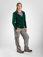 A woman modeling a dark green zip-up fleece jacket, paired with light gray cargo pants and hiking boots, against a light gray studio background.