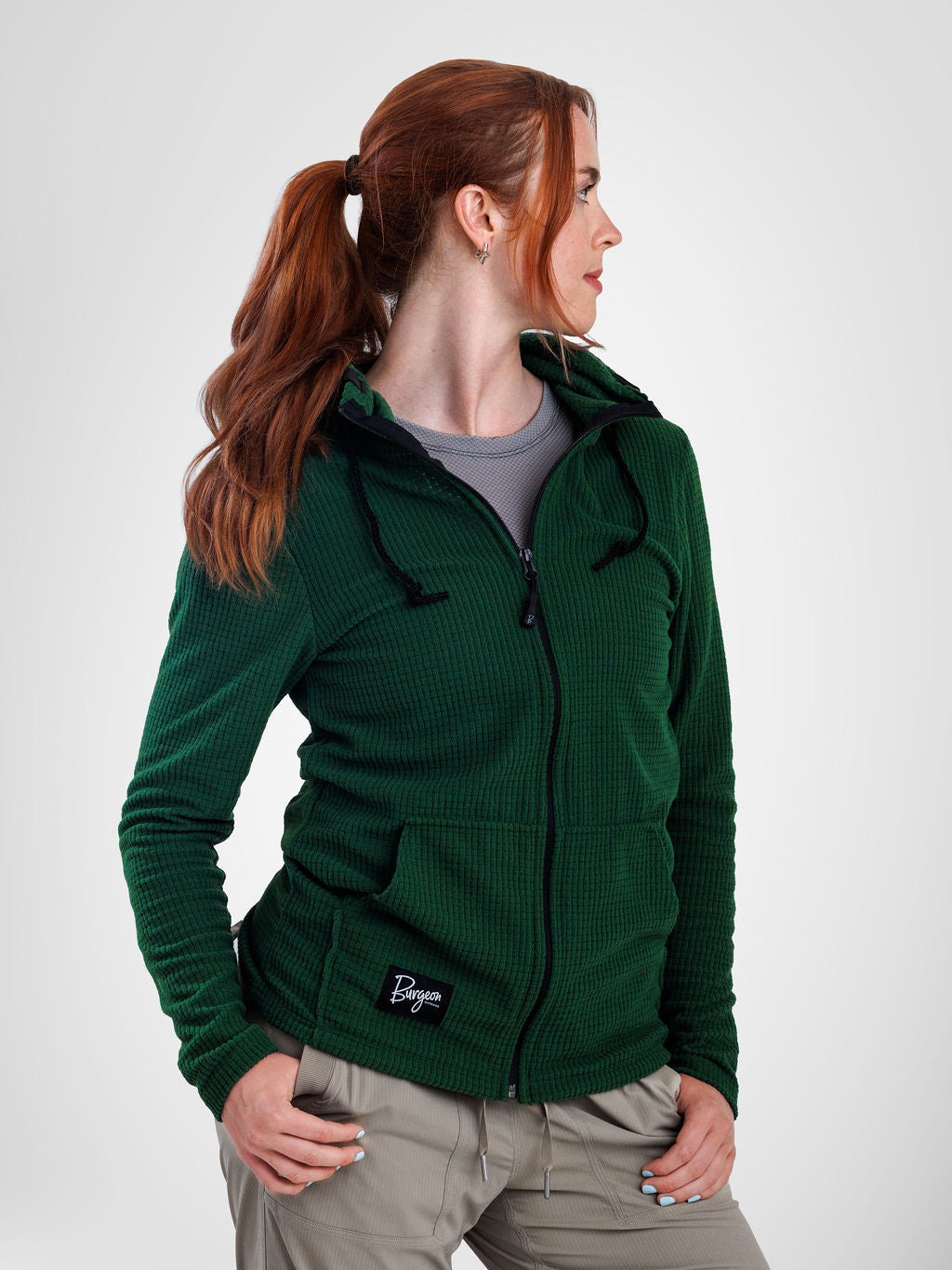 A woman with red hair in a ponytail wearing a dark green waffle-knit zip-up hoodie with a black logo patch on the front pocket, paired with a gray t-shirt and khaki pants, photographed against a light gray studio background.