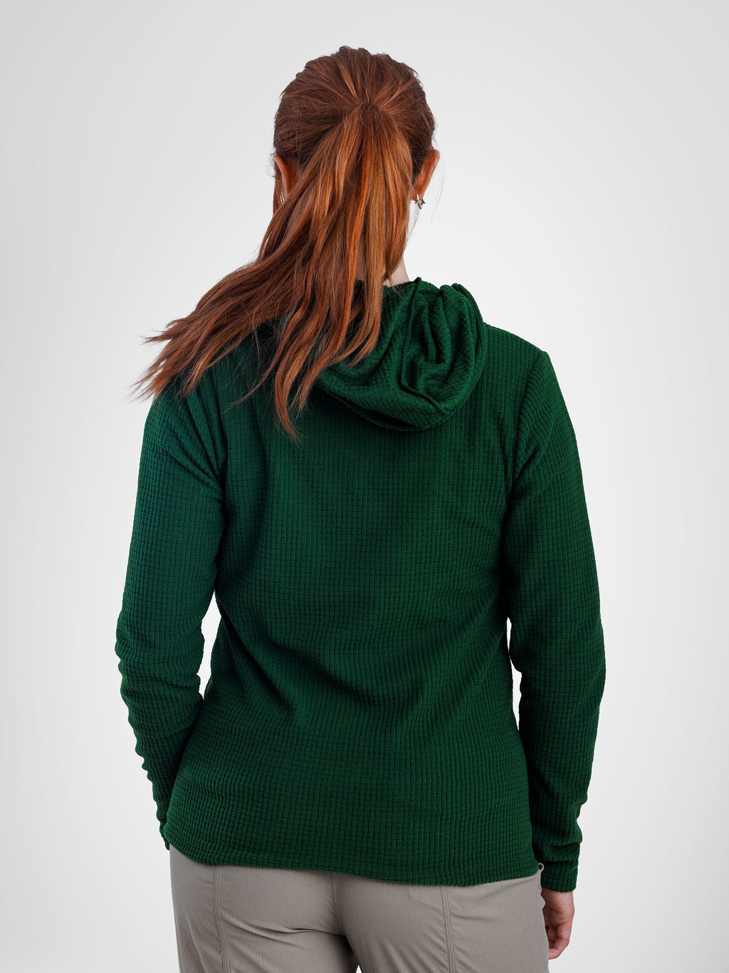 A woman viewed from behind wearing a dark green waffle-knit hooded long-sleeve top and light gray pants, against a plain light gray studio background.