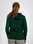 A woman viewed from behind wearing a dark green waffle-knit hooded long-sleeve top and light gray pants, against a plain light gray studio background.
