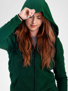 A woman wearing a dark green waffle-knit zip-up hoodie with the hood up, shown against a plain light gray studio background.