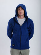 A man wearing a royal blue ribbed zip-up hoodie with a black zipper and a small black brand patch on the lower left hem, paired with a light gray t-shirt and khaki pants, against a plain light gray background.