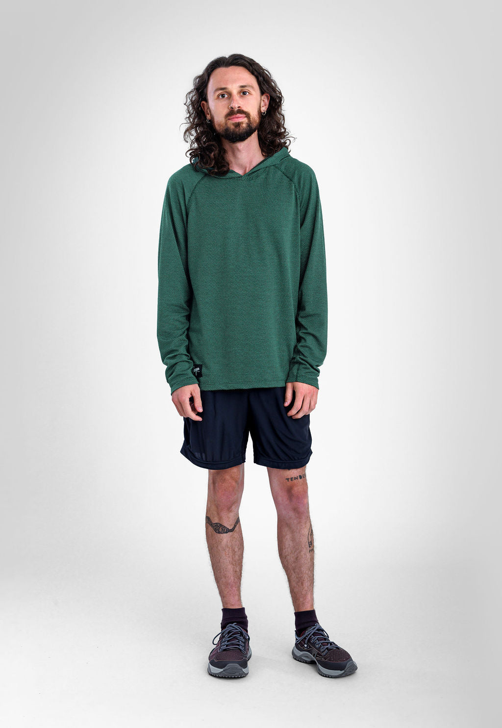 A man with long curly hair and a beard stands against a light gray studio background wearing a dark green long-sleeve Sunseeker Hoodie, dark navy athletic shorts, dark socks, and gray running shoes.