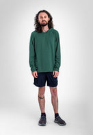 A man with long curly hair and a beard stands against a light gray studio background wearing a dark green long-sleeve Sunseeker Hoodie, dark navy athletic shorts, dark socks, and gray running shoes.