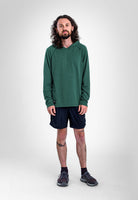 A man with long curly hair and a beard stands against a light gray studio background wearing a dark green long-sleeve Sunseeker Hoodie, dark navy athletic shorts, dark socks, and gray running shoes.