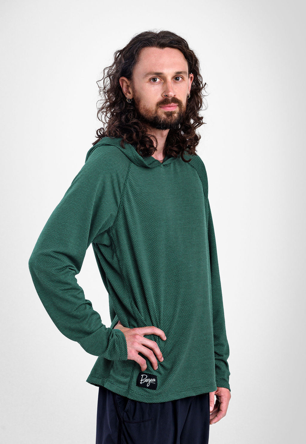 A man with long curly brown hair and a beard wearing a dark green long-sleeve Sunseeker Hoodie with a small black logo patch on the hem, posed against a light gray studio background.