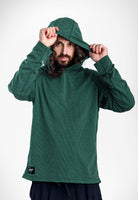 A man wearing a dark green Sunseeker Hoodie with a textured knit pattern, holding up the hood with both hands against a plain white studio background.