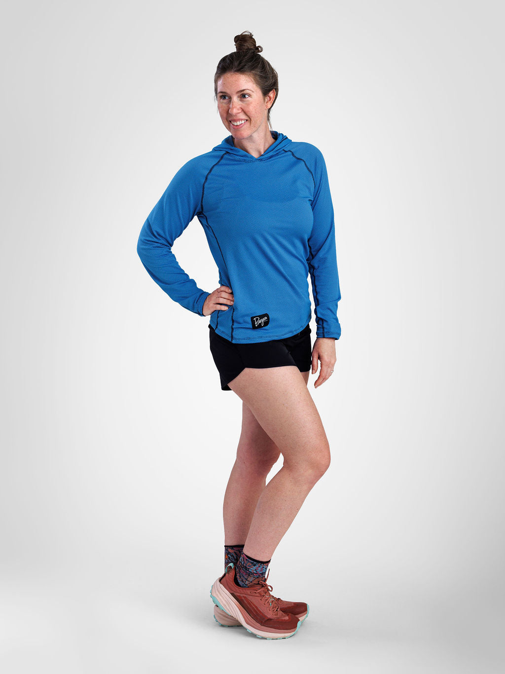 A woman modeling a bright blue long-sleeve sun hoodie with black running shorts, patterned socks, and brown trail running shoes against a light gray studio background.