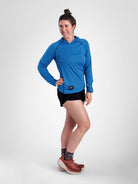 A woman modeling a bright blue long-sleeve sun hoodie with black running shorts, patterned socks, and brown trail running shoes against a light gray studio background.