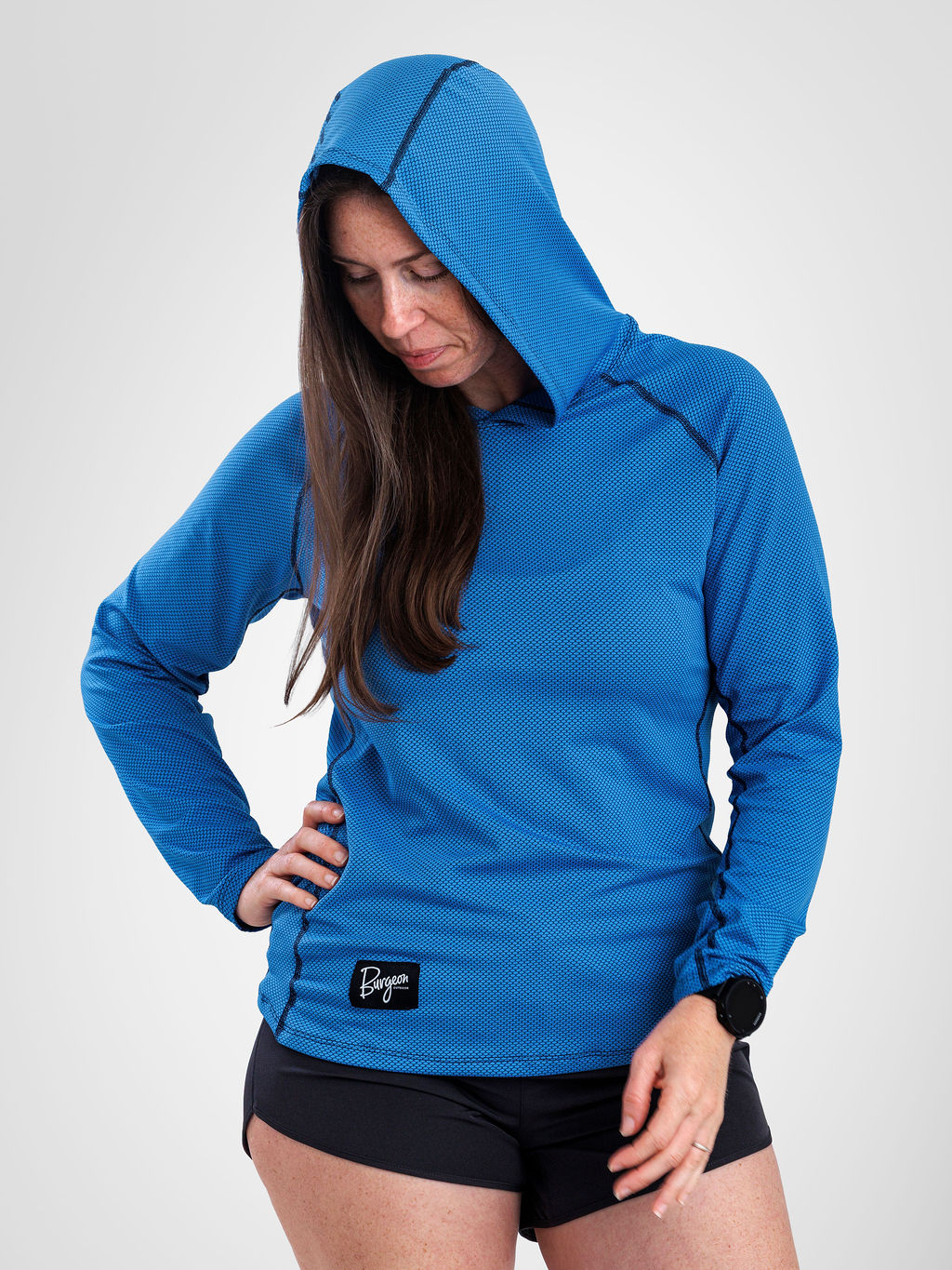 A woman wearing a bright blue long-sleeve sun hoodie with a small black logo patch on the front hem, paired with black athletic shorts, photographed against a light gray studio background.