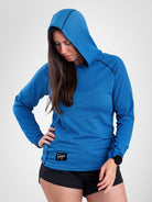 A woman wearing a bright blue long-sleeve sun hoodie with a small black logo patch on the front hem, paired with black athletic shorts, photographed against a light gray studio background.