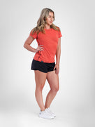 A woman modeling a coral orange short-sleeve athletic t-shirt and black athletic shorts, paired with white sneakers, against a light gray studio background.