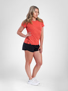 A woman modeling a coral orange short-sleeve athletic t-shirt and black athletic shorts, paired with white sneakers, against a light gray studio background.