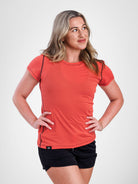 A woman modeling a fitted short-sleeve athletic top in a coral-orange color with thin black piping details along the shoulders and sides, paired with black shorts, against a light gray studio background.