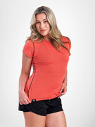 A woman modeling a fitted short-sleeve athletic top in a coral-orange color with black side piping, paired with black athletic shorts, against a plain light gray studio background.