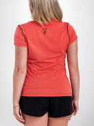 A woman viewed from the back wearing a fitted short-sleeve athletic top in a coral-orange color with black piping along the shoulders and a small black logo near the upper back, paired with black shorts against a light gray studio background.
