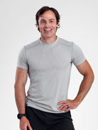 A smiling man wearing a light gray short-sleeve athletic t-shirt and black pants, standing against a light gray studio background.