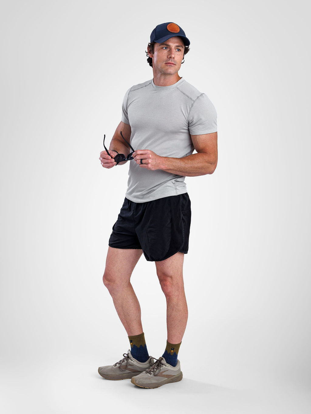 A man modeling a light gray short-sleeve athletic t-shirt, black athletic shorts, a navy blue baseball cap with an orange logo, olive green socks, and gray trail running shoes against a light gray studio background.
