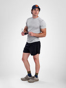 A man modeling a light gray short-sleeve athletic t-shirt, black athletic shorts, a navy blue baseball cap with an orange logo, olive green socks, and gray trail running shoes against a light gray studio background.