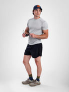 A man modeling a light gray short-sleeve athletic t-shirt, black athletic shorts, a navy blue baseball cap with an orange logo, olive green socks, and gray trail running shoes against a light gray studio background.