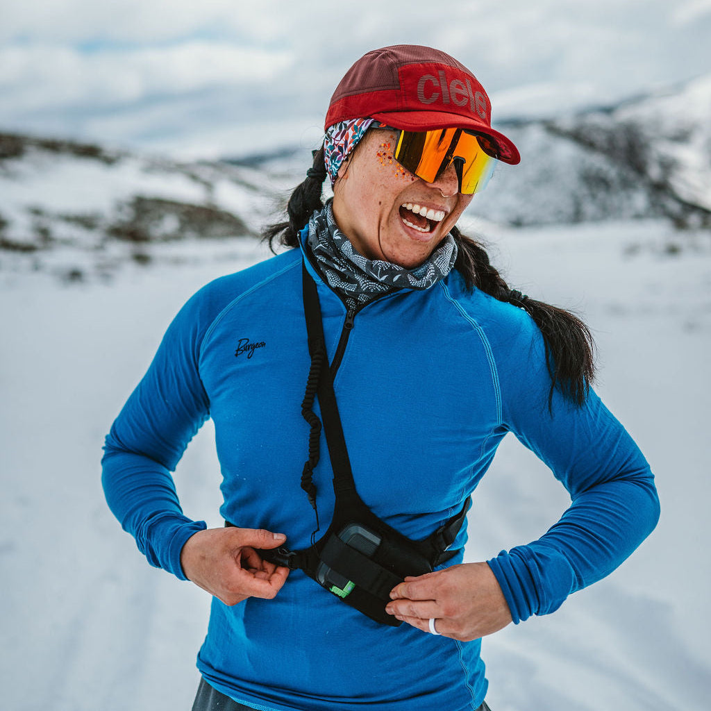 A woman in a snowy outdoor setting wearing a bright blue long-sleeve athletic top, a red cap, reflective orange-tinted goggles, and a black chest-mounted storage pack.